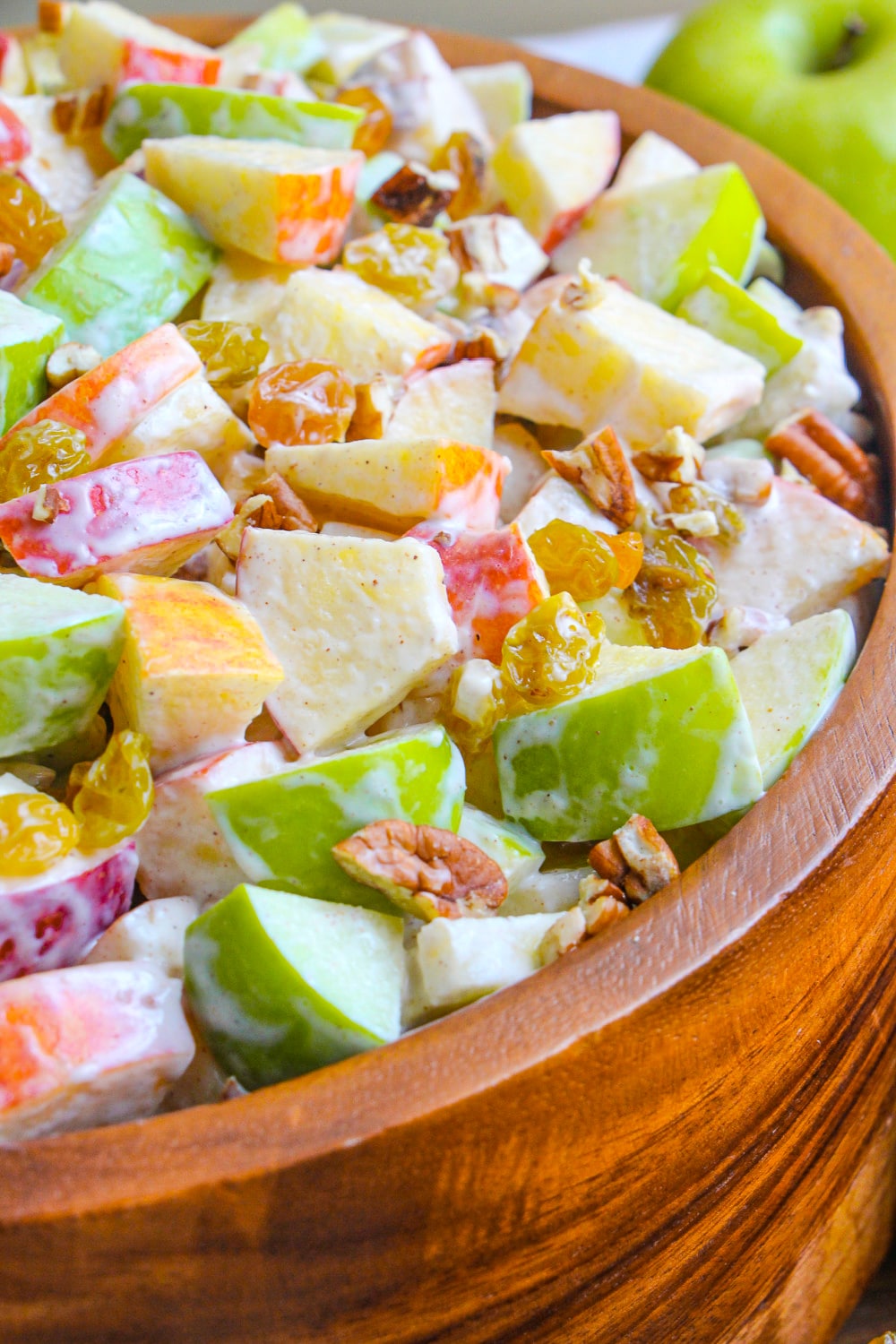 Chopped red and green apples tossed in cream cheese cinnamon dressing garnished with golden raisins and pecans in a larger wood serving bowl.