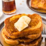 A stack of French Toast drenched in syrup and topped with butter sit on a plate next to a fork.