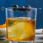 An Old Fashioned in a clear glass with a pick of cherries resting across the rim.