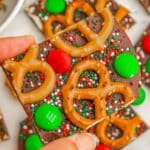 A woman's hand holding a piece of chocolate bark with mini pretzels, M&M candies, and red and green sprinkles.