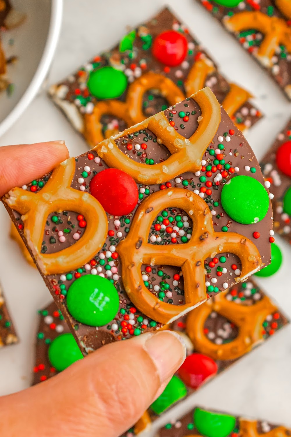 A woman's hand holding a piece of chocolate bark with mini pretzels, M&M candies, and red and green sprinkles.