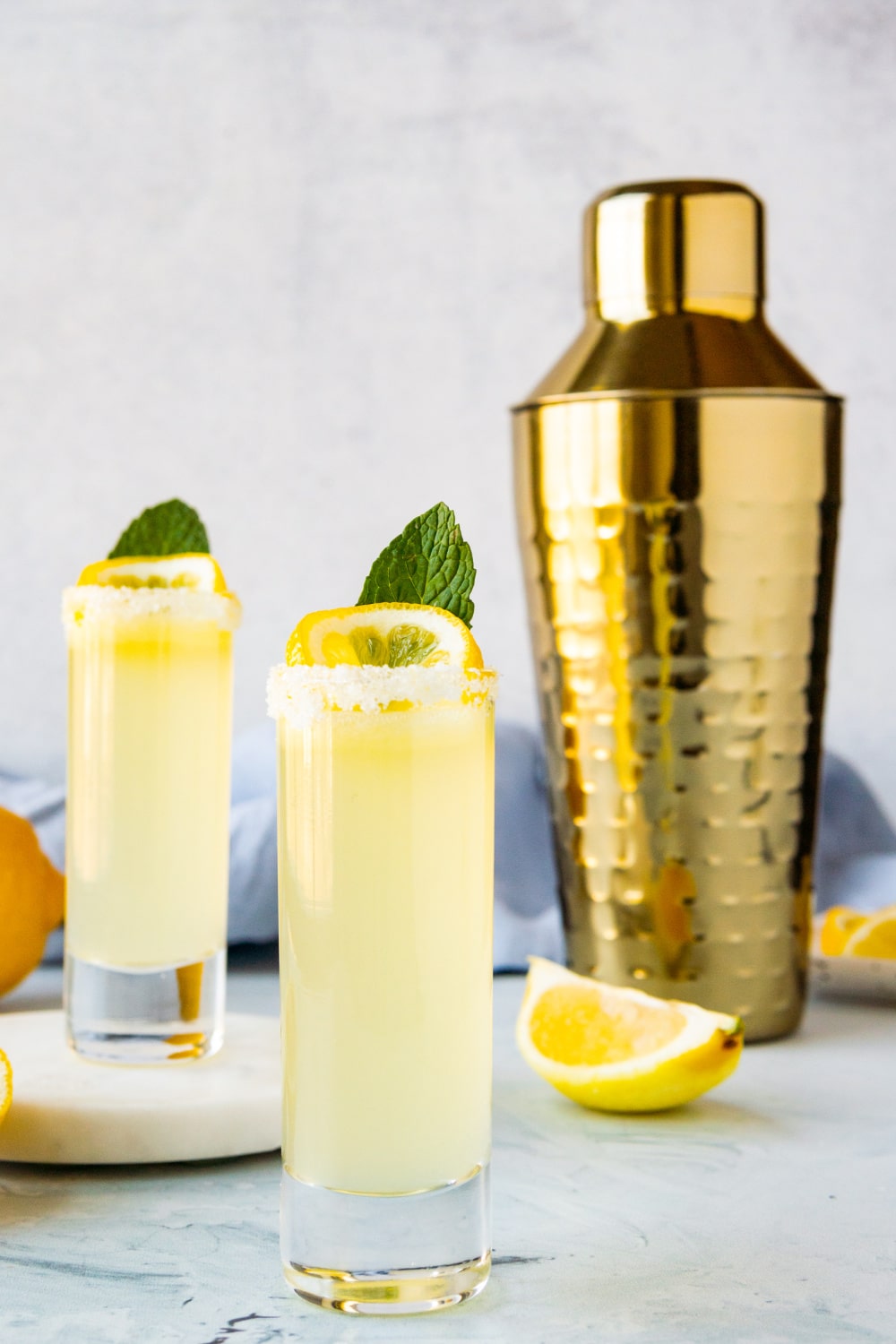 Two Lemon Drop Shots sit next to a cocktail shaker.