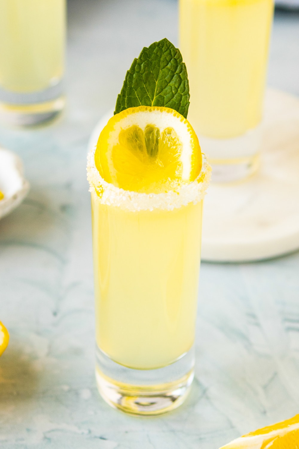 A single shot garnished with a lemon slice and a mint leaf sits on a marble countertop.