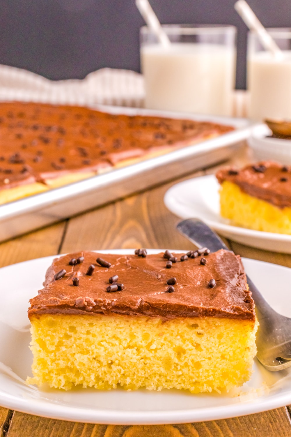 A slice of Yellow Sheet Cake with Chocolate Frosting on a white dessert plate sits in front of the remaining cake in the sheet pan.