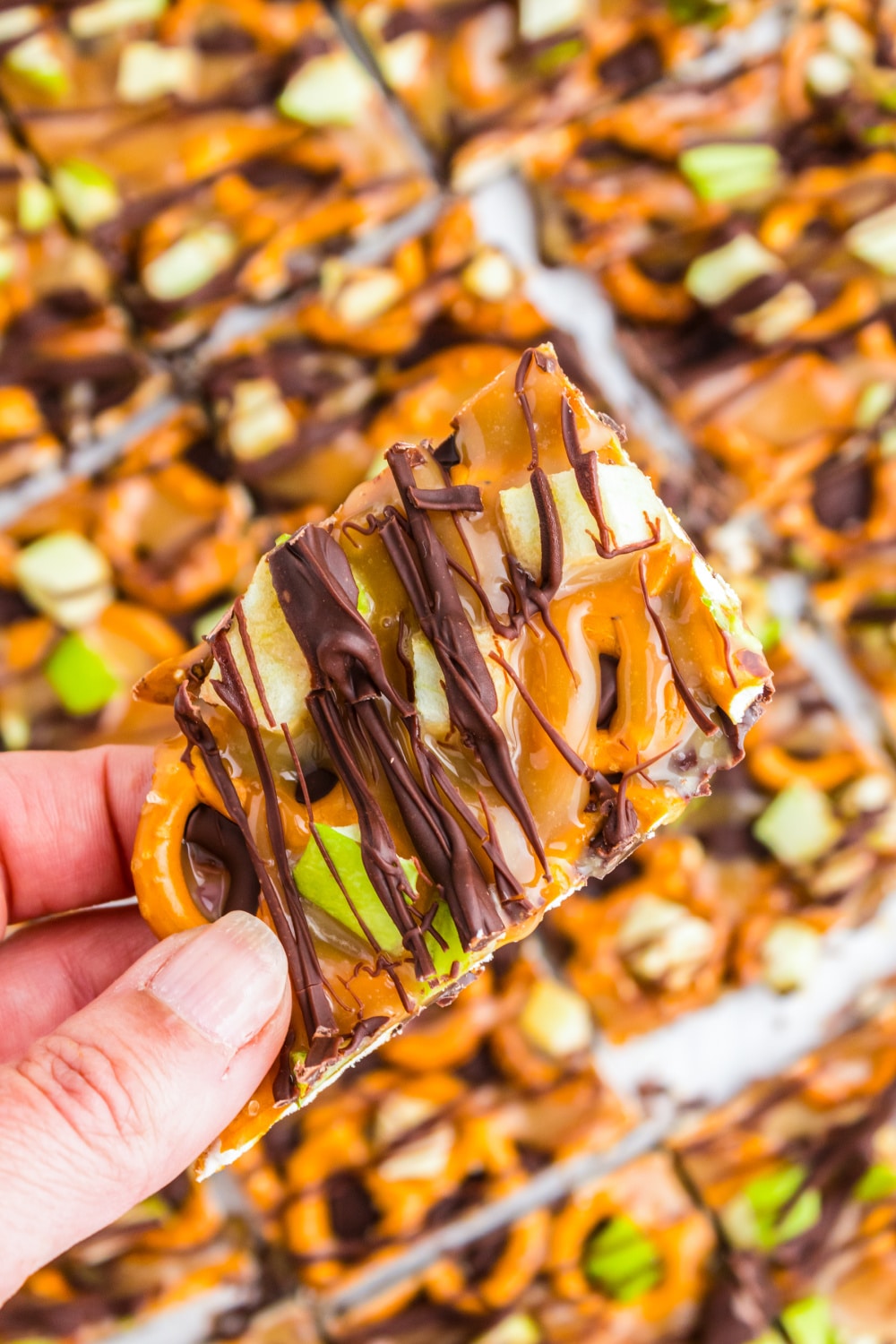A single piece of Caramel Apple Bark is held in a woman's hand over a tray of pieces of the same.