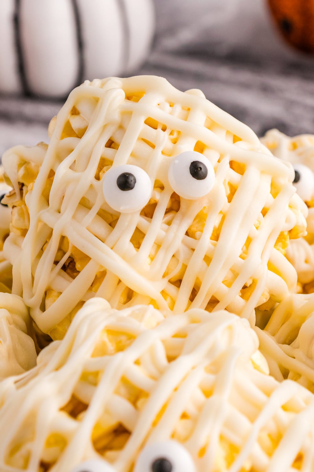 A popcorn ball drizzled in white chocolate to resemble a mummy sits on a plate with other popcorn balls.