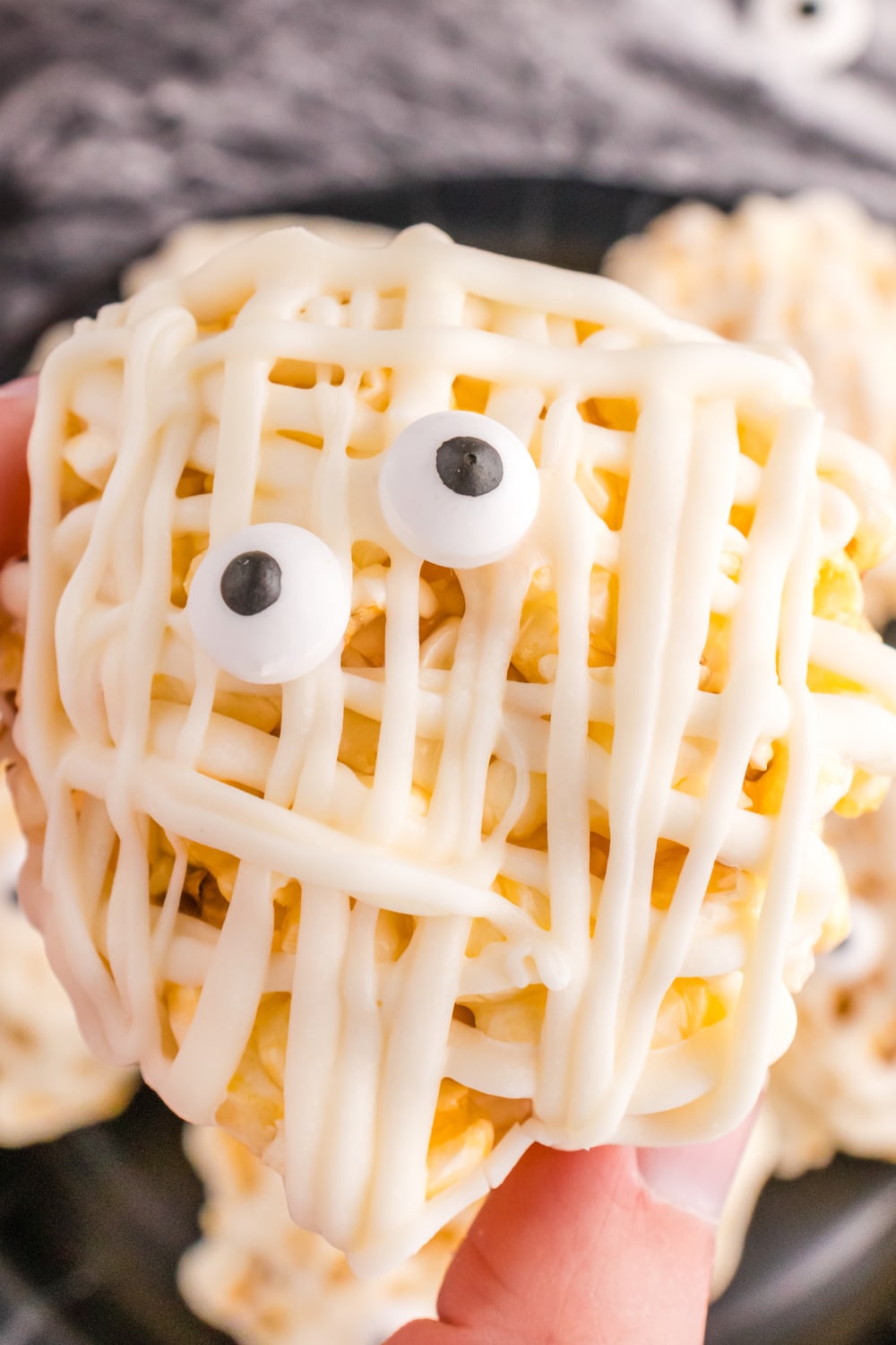 A woman's hand holds a single popcorn ball drizzled in white chocolate with a pair of candy eyes.