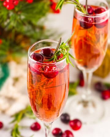 Two champagne glasses with mimosas garnished with rosemary and fresh cranberry.