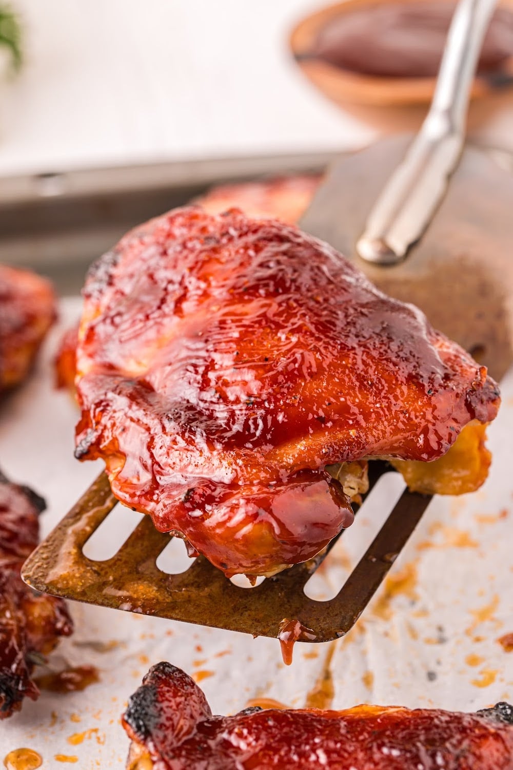 A single piece of chicken on a spatula over a sheet pan filled of BBQ Chicken.