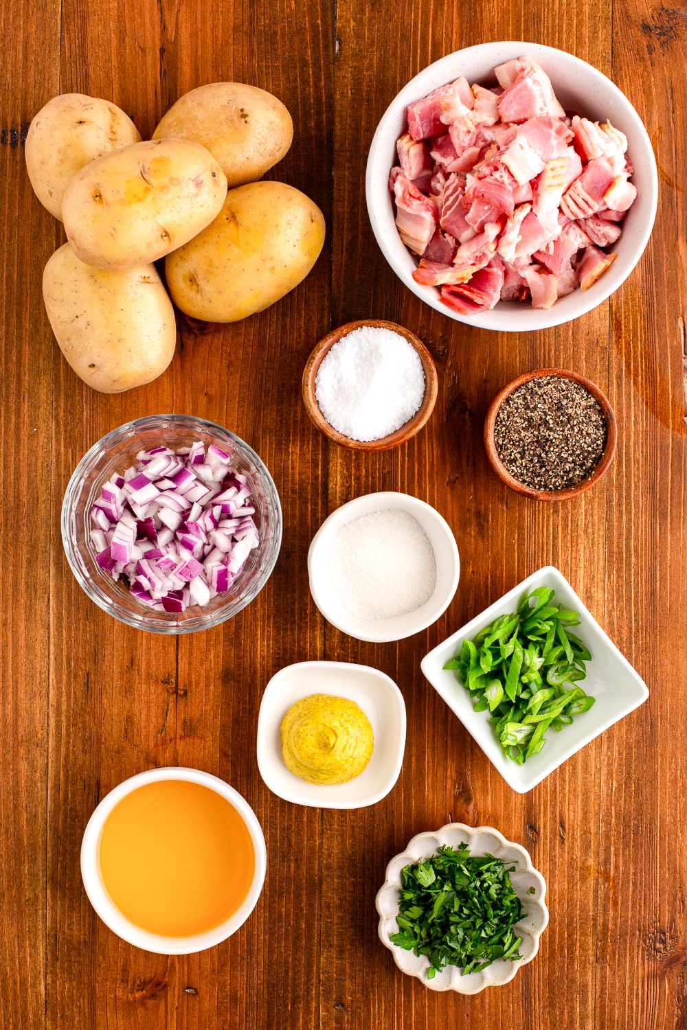 Measured ingredients need to make this potato salad recipe presented in small bowls on a wood cutting board.