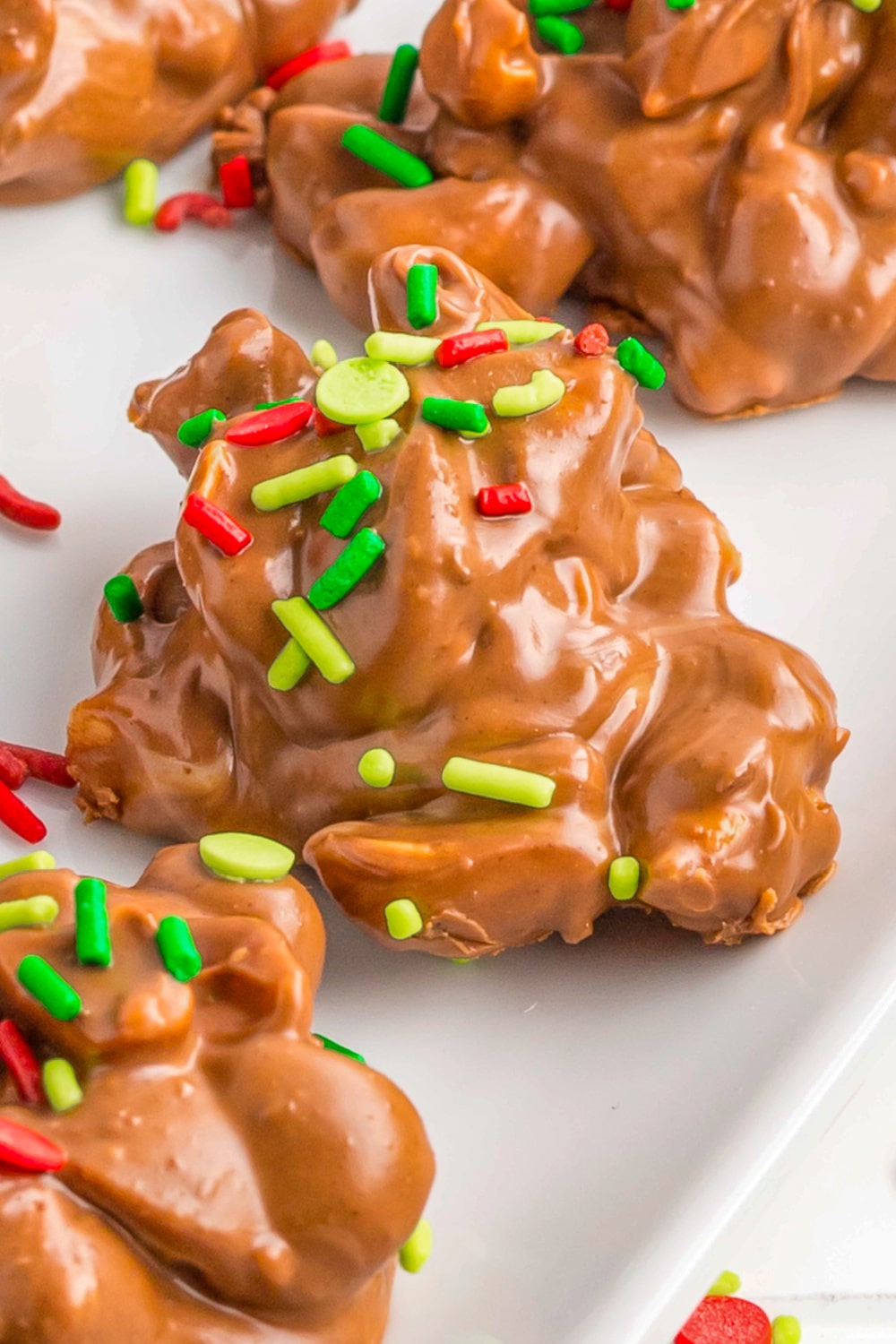 Red and green sprinkles top chocolate peanut candy string on a white plate.